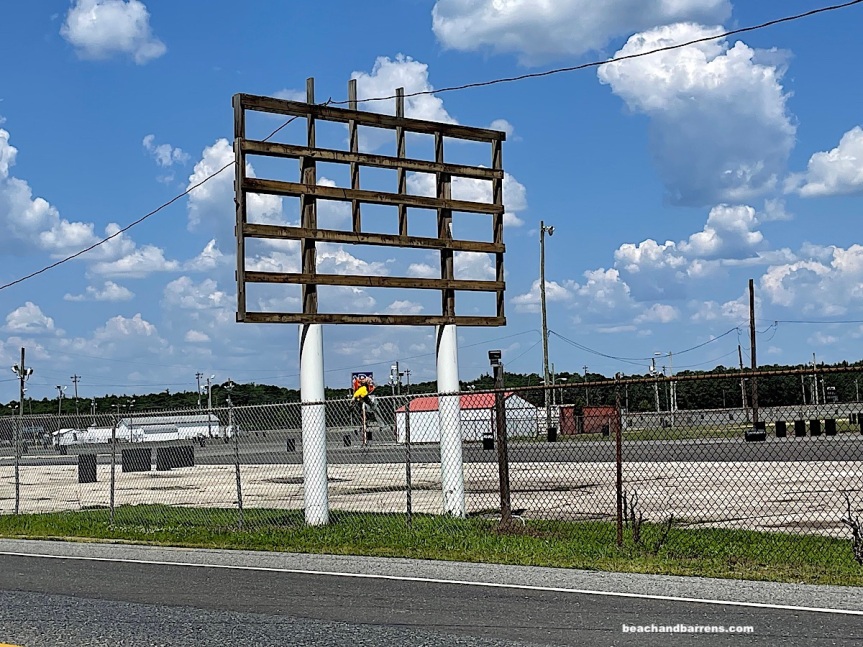 Atco racetrack behind chainlink fence adorned with yellow bouquet of flowers underneath signpost with sign removed, border of green grass and road in foreground, blue sky with puffy white clouds in background: beachandbarrens.com watermark