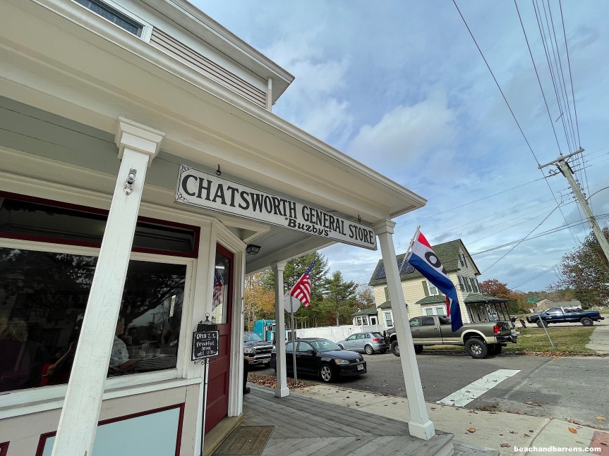 White columns and sign reading "Chatsworth General Store - 'Buzby's" with wooden front steps and maroon door on street in Chatsworth with American flag and Open flag, ; beachandbarrens.com watermark