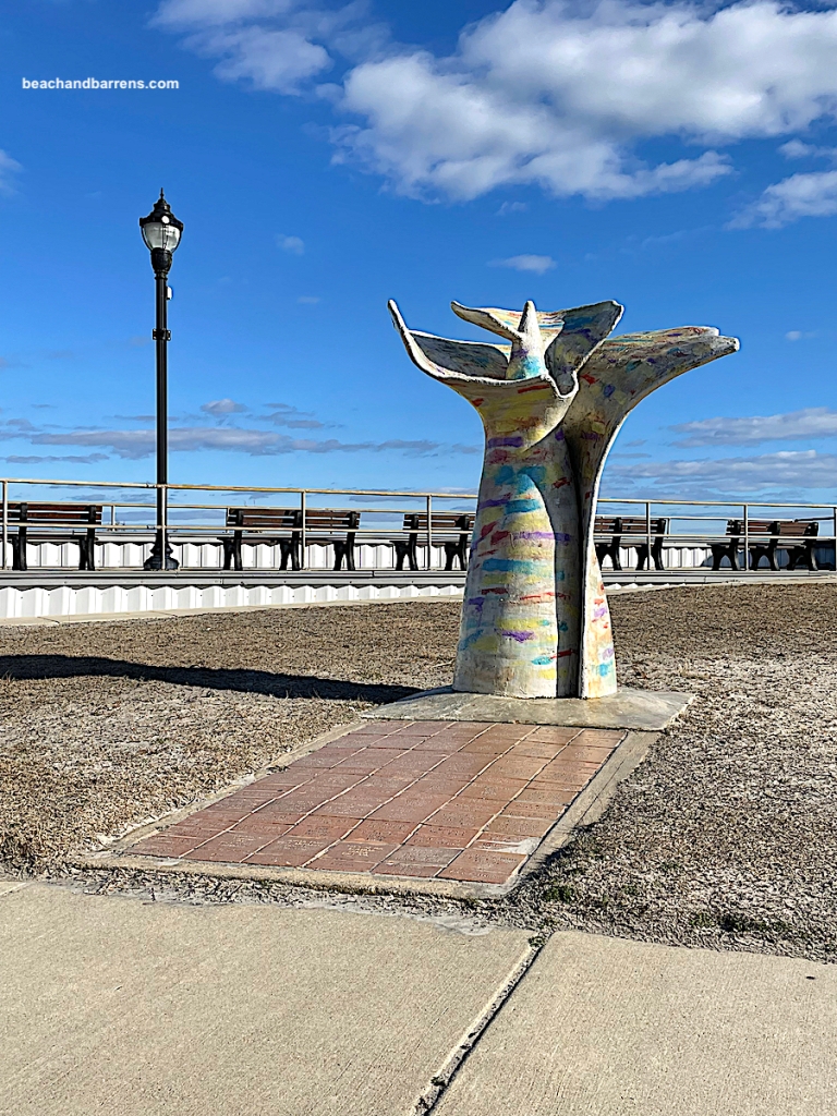 Alabaster multi-colored public seashell sculpture called Spirit of Brigantine by Gregg Knight of Knight Sculpture Studios with the silver railing, benches, and black lamp post of the Brigantine Seawall in background under a blue sky with white clouds, beachandbarrens.com watermark in upper left corner